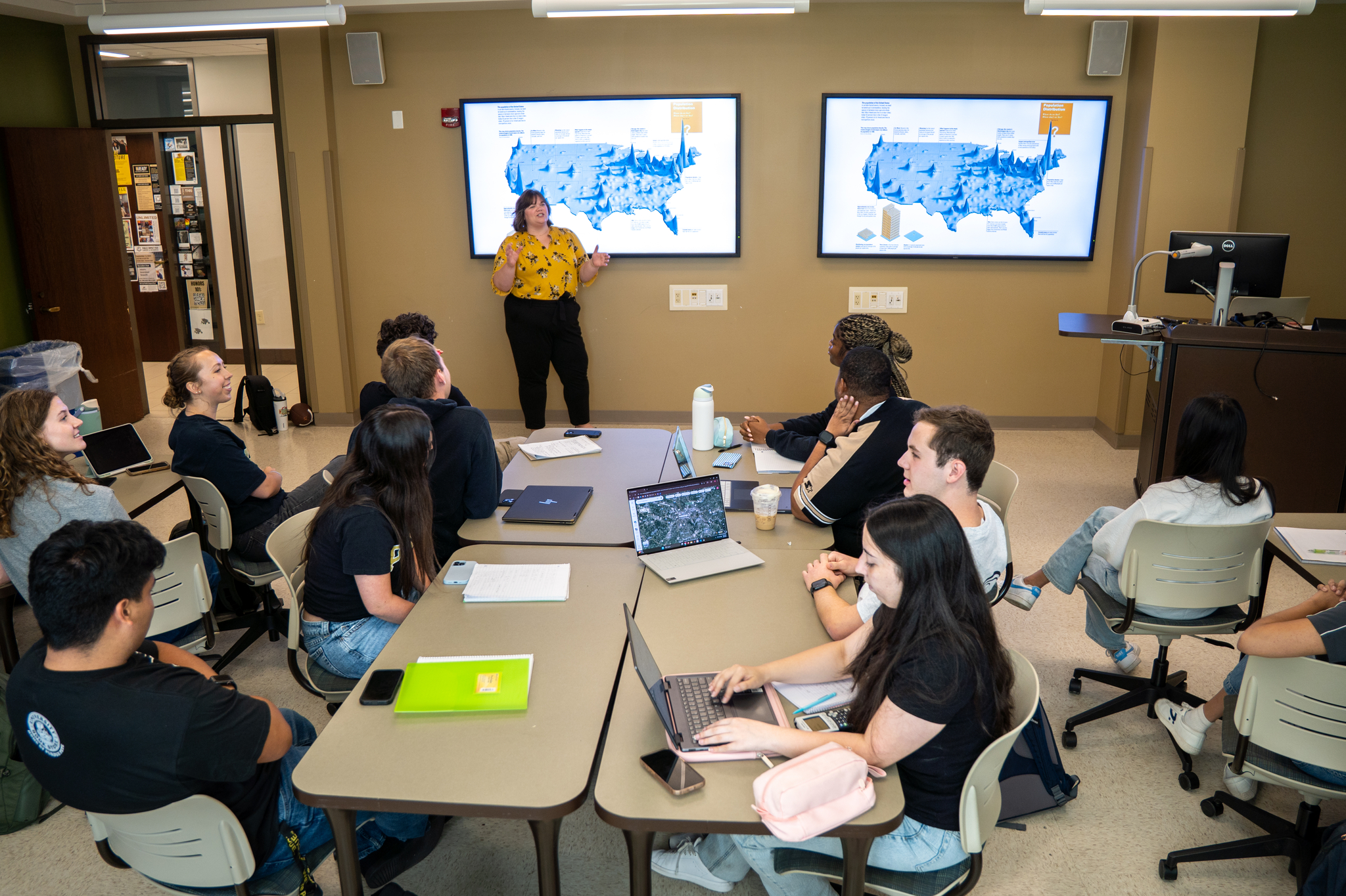 Students in a small classroom looking at a woman presenting in front of two TV screens.